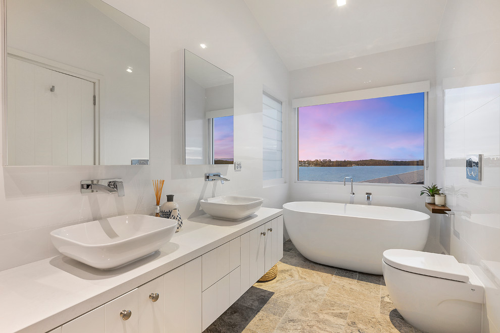 Inspiration for a contemporary 3/4 bathroom in Central Coast with flat-panel cabinets, white cabinets, a freestanding tub, a wall-mount toilet, white tile, white walls, a vessel sink and beige floor.