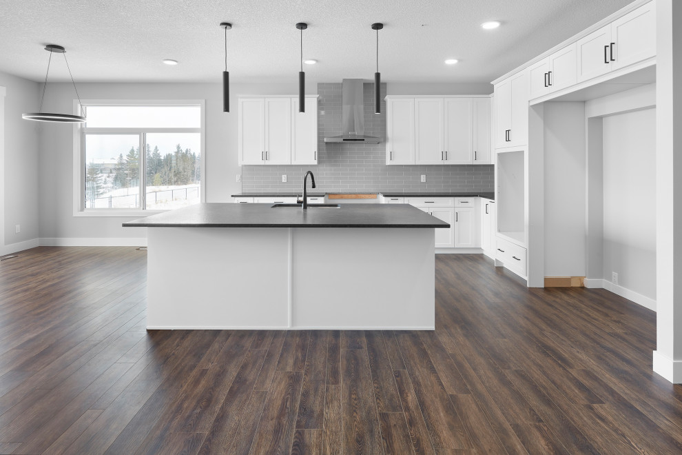 Design ideas for a mid-sized modern l-shaped open plan kitchen in Edmonton with an undermount sink, shaker cabinets, white cabinets, granite benchtops, grey splashback, subway tile splashback, stainless steel appliances, dark hardwood floors, with island, brown floor and black benchtop.