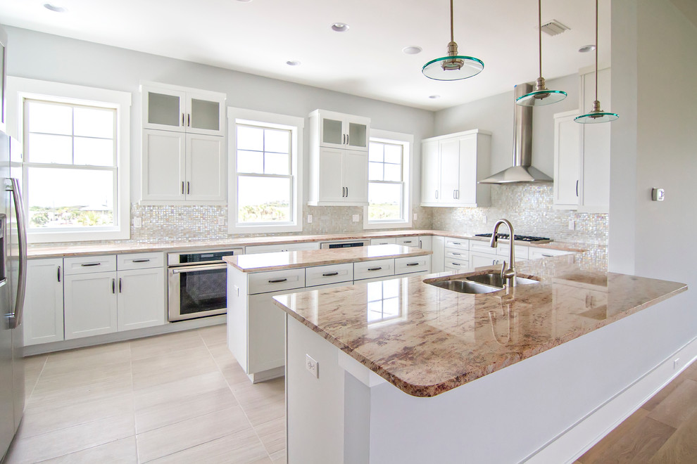 Large beach style u-shaped open plan kitchen in Jacksonville with a double-bowl sink, shaker cabinets, white cabinets, granite benchtops, white splashback, glass tile splashback, stainless steel appliances, porcelain floors, with island, beige floor and brown benchtop.