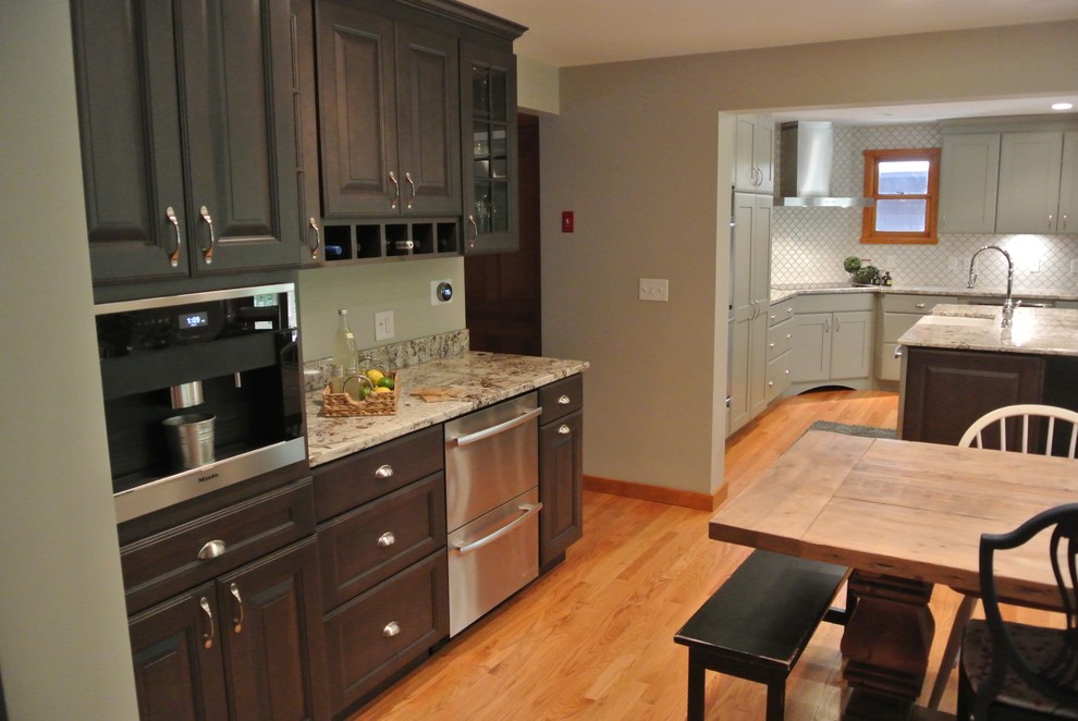 Example of a large transitional l-shaped light wood floor eat-in kitchen design in Boston with a farmhouse sink, shaker cabinets, gray cabinets, granite countertops, white backsplash, ceramic backsplash, stainless steel appliances, an island and gray countertops