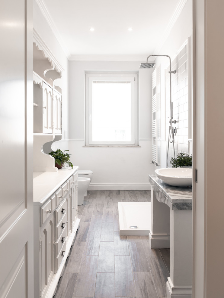 This is an example of a mid-sized contemporary master bathroom in Florence with recessed-panel cabinets, white cabinets, an open shower, white tile, ceramic tile, white walls, porcelain floors, a vessel sink, marble benchtops, beige floor, an open shower, grey benchtops, a single vanity, a freestanding vanity and a wall-mount toilet.