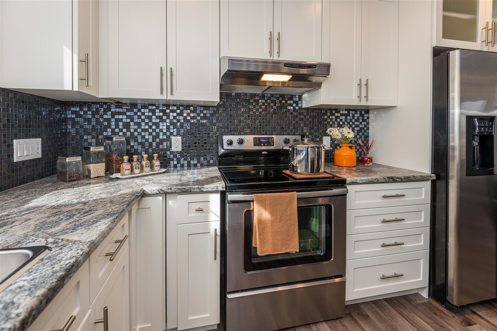 Example of a mid-sized transitional l-shaped medium tone wood floor and brown floor eat-in kitchen design with a drop-in sink, shaker cabinets, white cabinets, granite countertops, metallic backsplash, mosaic tile backsplash, stainless steel appliances, an island and multicolored countertops