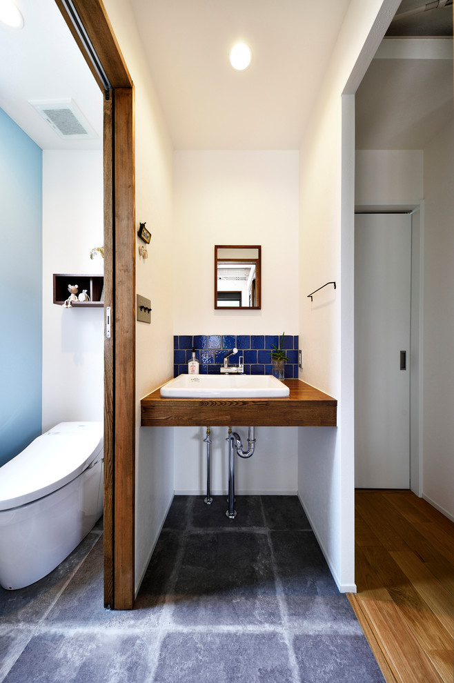Photo of an industrial powder room with blue tile, white walls, concrete floors, a console sink, grey floor, wood benchtops and brown benchtops.