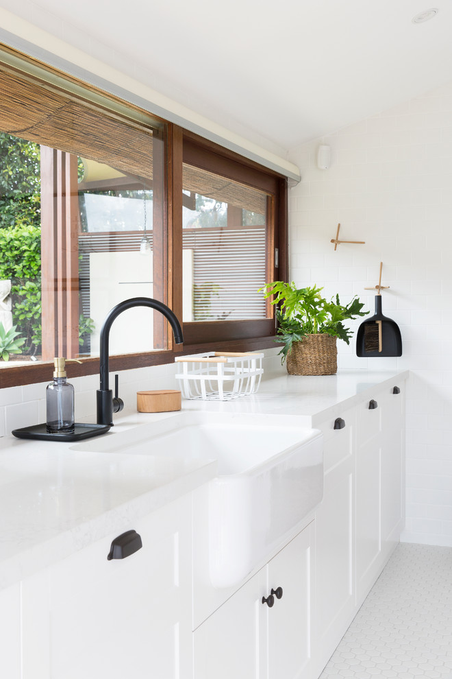 Inspiration for an expansive contemporary galley dedicated laundry room in Gold Coast - Tweed with a farmhouse sink, shaker cabinets, white cabinets, quartz benchtops, white walls, porcelain floors, a stacked washer and dryer and white floor.