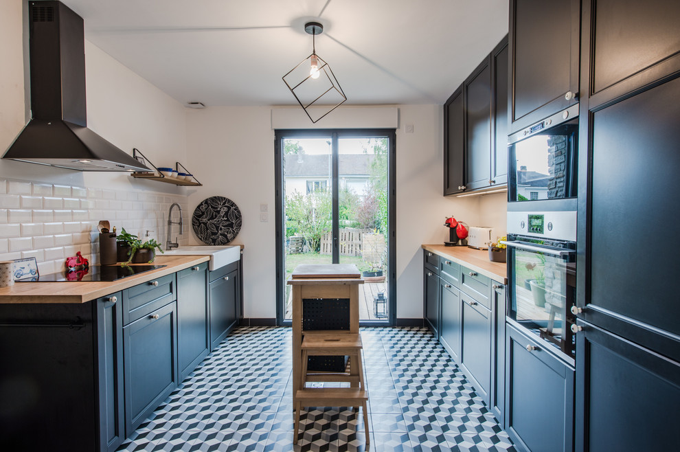 Photo of a scandinavian galley separate kitchen in Montpellier with a farmhouse sink, recessed-panel cabinets, black cabinets, wood benchtops, white splashback, subway tile splashback, stainless steel appliances, with island, multi-coloured floor and beige benchtop.