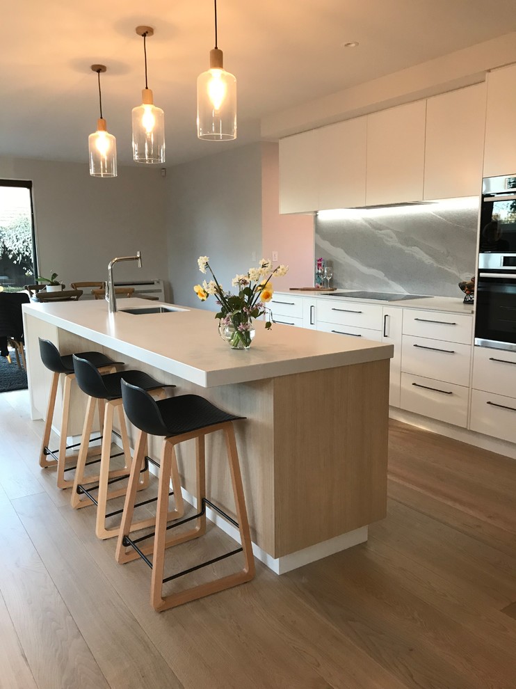 Design ideas for a large contemporary galley open plan kitchen in Christchurch with a single-bowl sink, flat-panel cabinets, white cabinets, quartz benchtops, grey splashback, stone slab splashback, stainless steel appliances, medium hardwood floors, with island, brown floor and white benchtop.