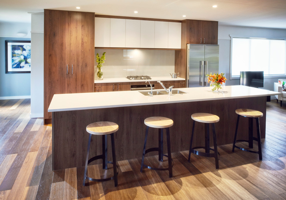 Photo of a mid-sized beach style galley kitchen in Auckland with a double-bowl sink, flat-panel cabinets, medium wood cabinets, quartz benchtops, grey splashback, glass sheet splashback, stainless steel appliances, medium hardwood floors and with island.