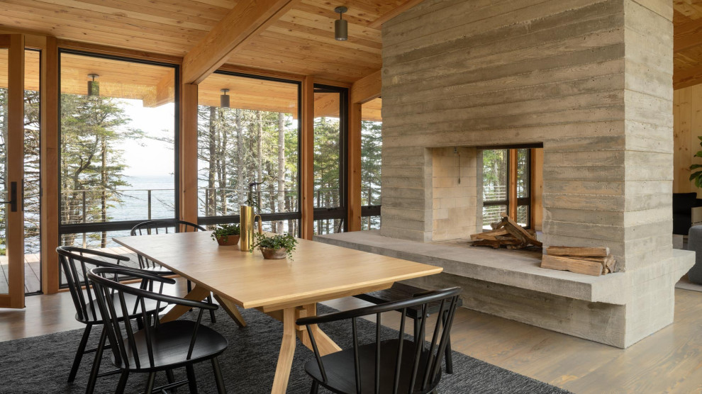 This is an example of a mid-sized country open plan dining in Portland Maine with brown walls, medium hardwood floors, a two-sided fireplace, a concrete fireplace surround and grey floor.