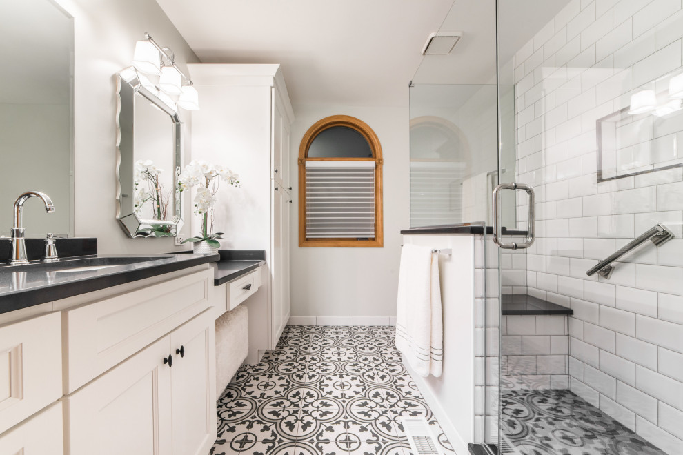 Transitional bathroom with recessed-panel cabinets, white cabinets, a corner shower, white tile, white walls, cement tiles, an undermount sink, multi-coloured floor, a hinged shower door and black benchtops.