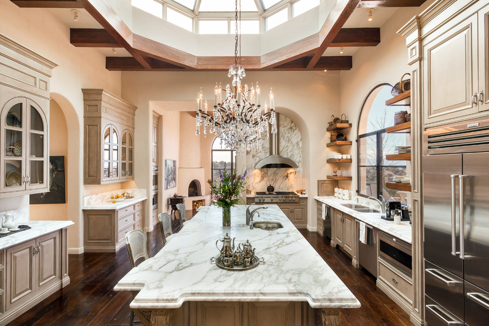 This is an example of a mediterranean l-shaped kitchen in Albuquerque with a double-bowl sink, raised-panel cabinets, beige cabinets, marble benchtops, stainless steel appliances, dark hardwood floors, with island and stone slab splashback.