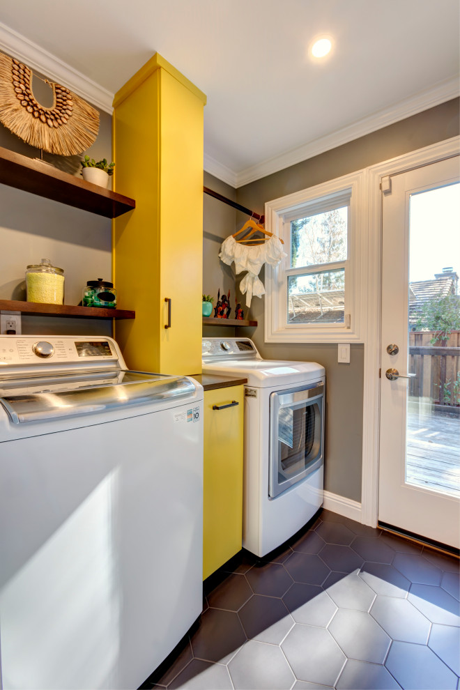 This is an example of a small eclectic laundry room in San Francisco with flat-panel cabinets, yellow cabinets, wood benchtops, grey walls, porcelain floors, a side-by-side washer and dryer and black floor.