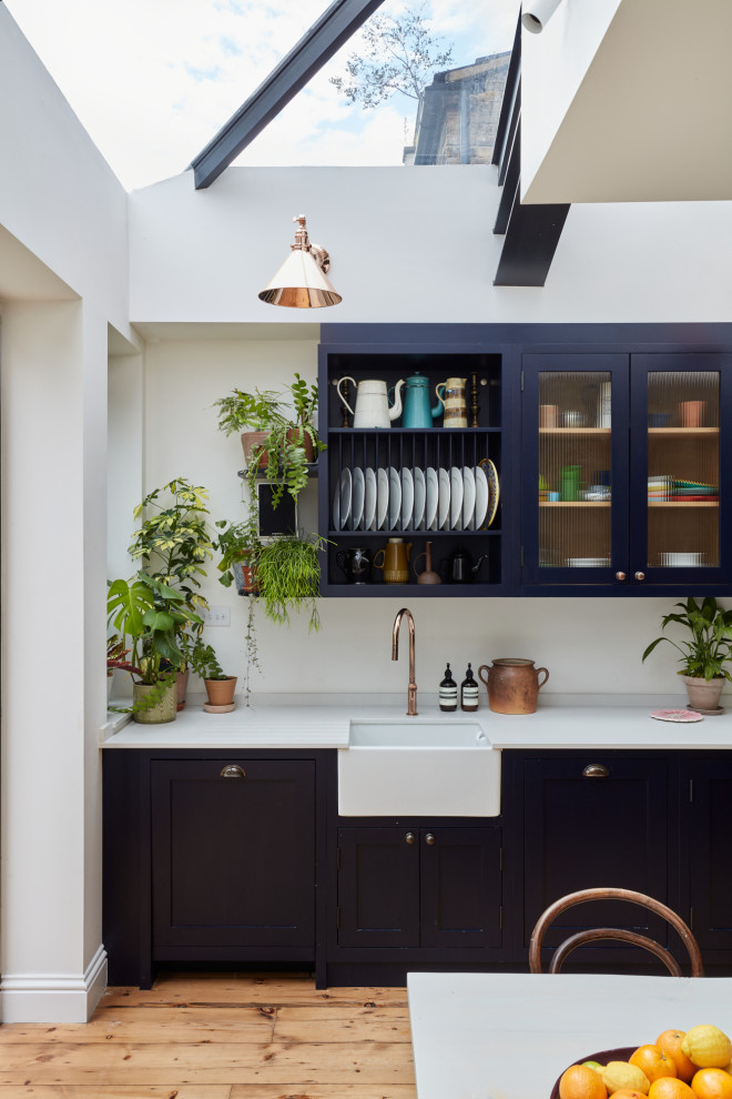 Photo of a large eclectic l-shaped eat-in kitchen in London with a farmhouse sink, shaker cabinets, blue cabinets, quartz benchtops, white splashback, engineered quartz splashback, black appliances, light hardwood floors, no island, grey benchtop and exposed beam.