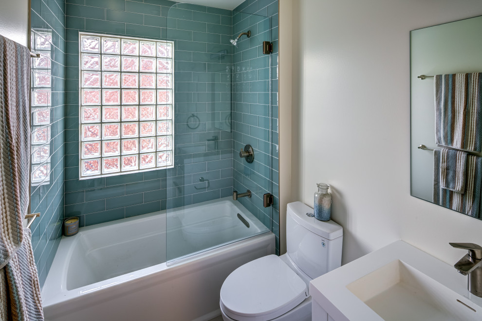 Photo of a mid-sized transitional kids bathroom in DC Metro with flat-panel cabinets, white cabinets, an alcove tub, a shower/bathtub combo, a one-piece toilet, blue tile, glass tile, white walls, ceramic floors, an integrated sink, solid surface benchtops, beige floor, a hinged shower door and white benchtops.