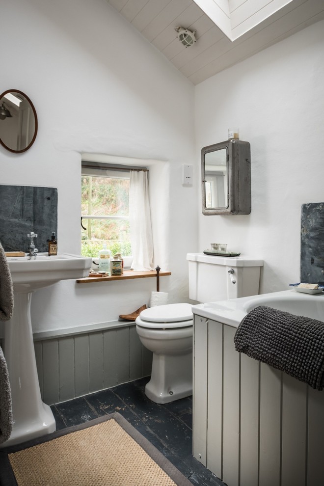 This is an example of a small country kids bathroom in Cornwall with shaker cabinets, grey cabinets, a drop-in tub, a shower/bathtub combo, a two-piece toilet, white walls, medium hardwood floors, a pedestal sink and black floor.