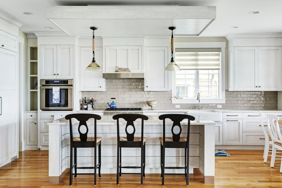 This is an example of a beach style l-shaped eat-in kitchen in Other with a farmhouse sink, shaker cabinets, white cabinets, beige splashback, subway tile splashback, panelled appliances, medium hardwood floors, with island, brown floor and beige benchtop.