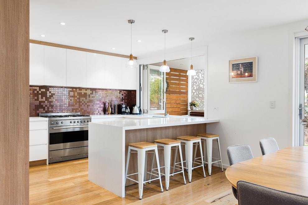 Photo of a mid-sized contemporary u-shaped eat-in kitchen in Sydney with an undermount sink, white cabinets, metallic splashback, mosaic tile splashback, stainless steel appliances, light hardwood floors, solid surface benchtops, with island, brown floor and white benchtop.
