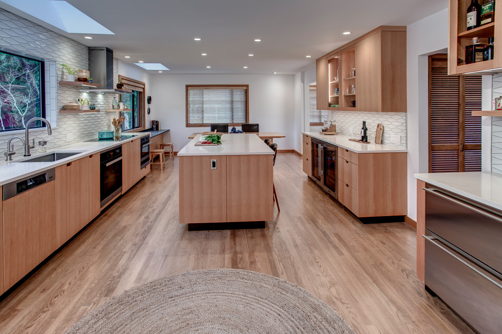 This is an example of a large modern kitchen in Seattle with an undermount sink, flat-panel cabinets, light wood cabinets, quartz benchtops, white splashback, porcelain splashback, stainless steel appliances, light hardwood floors, with island and white benchtop.