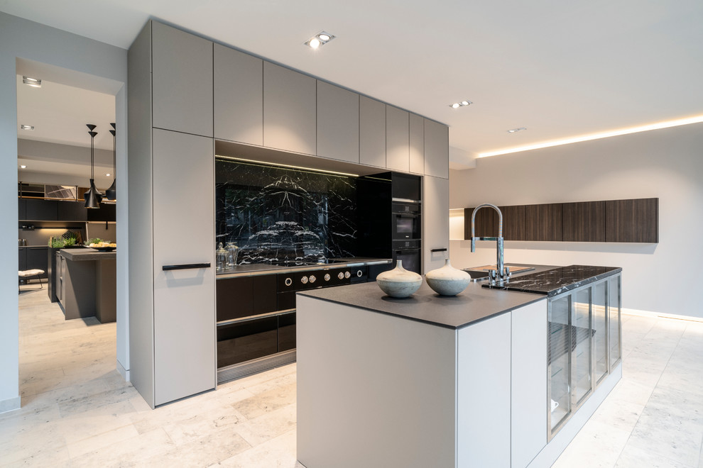 Photo of a large contemporary galley open plan kitchen in Cologne with a drop-in sink, flat-panel cabinets, grey cabinets, solid surface benchtops, grey splashback, black appliances, marble floors, with island, beige floor and black benchtop.