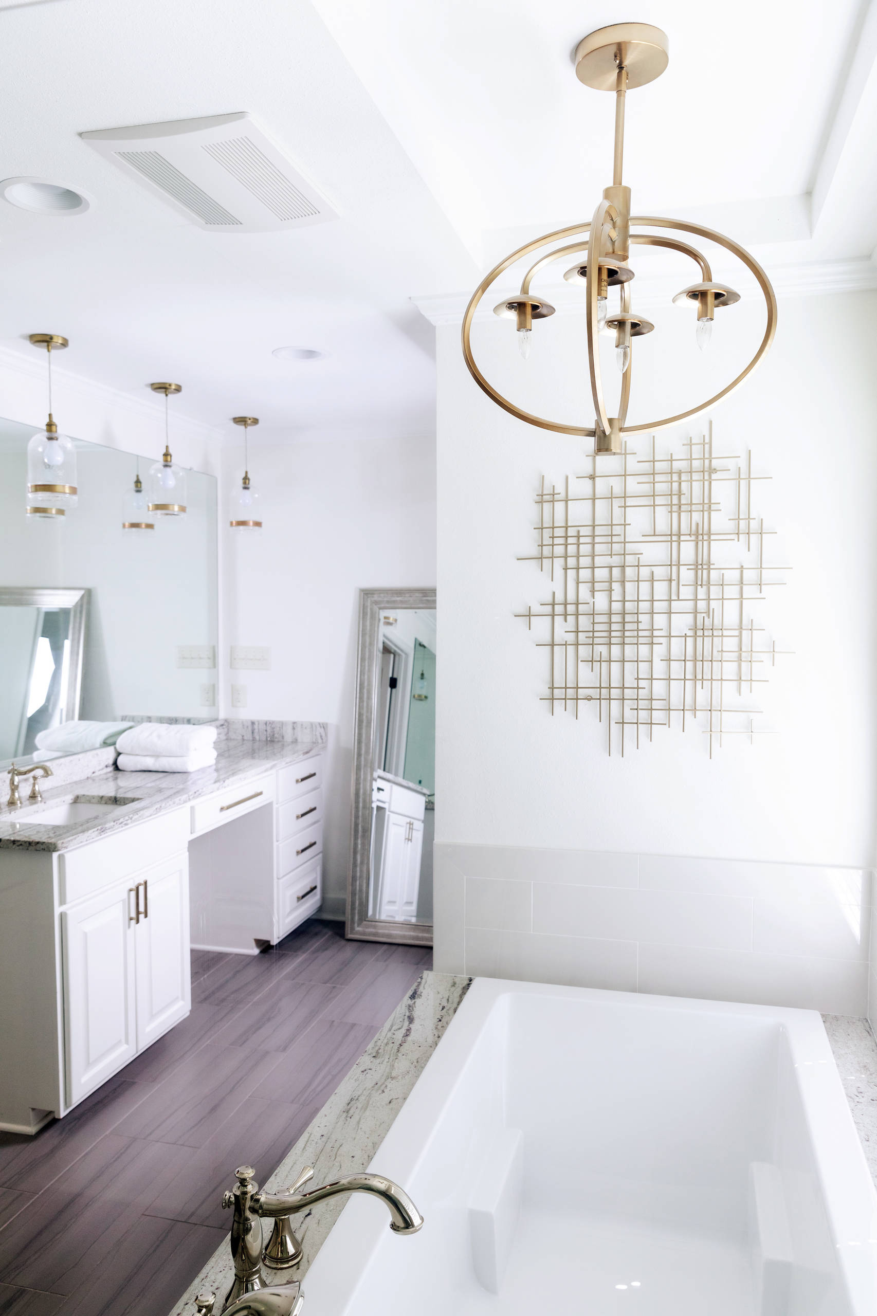 Vanity with white cabinets and honey bronze hardware. Frameless mirror above vanity. Glass pendant light with antique copper accents. Satin brass pendant light over tub. Framed mirror leaned up agains