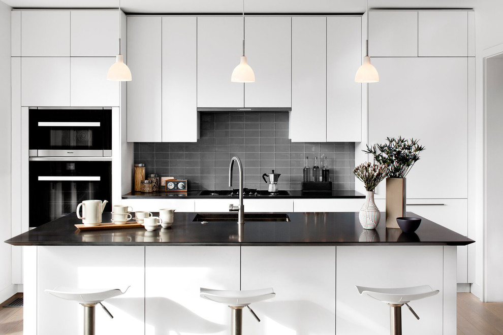 Design ideas for a modern kitchen in DC Metro with an undermount sink, flat-panel cabinets, quartz benchtops, grey splashback, light hardwood floors, with island, glass tile splashback and black appliances.