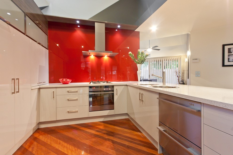 Small modern u-shaped eat-in kitchen in Melbourne with a double-bowl sink, quartz benchtops, red splashback, glass sheet splashback and stainless steel appliances.