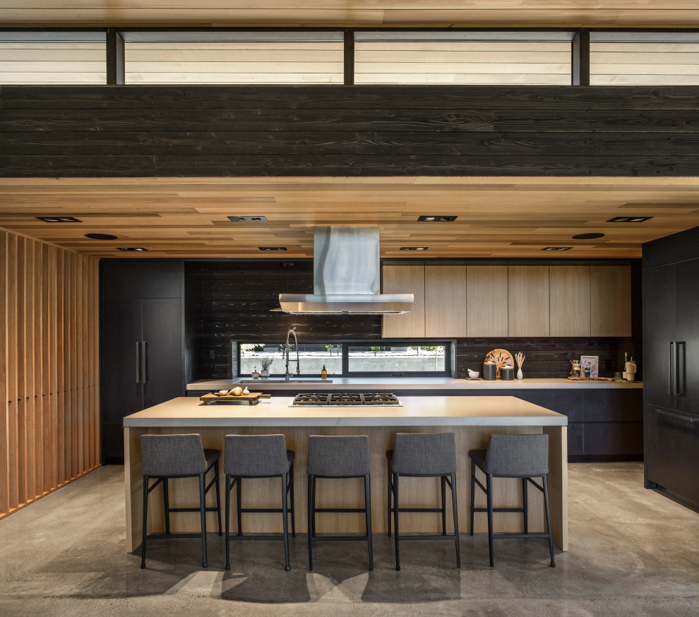 Modern kitchen in Portland with grey cabinets, concrete benchtops, glass sheet splashback, black appliances, with island, grey benchtop, an undermount sink, concrete floors, grey floor and wood.