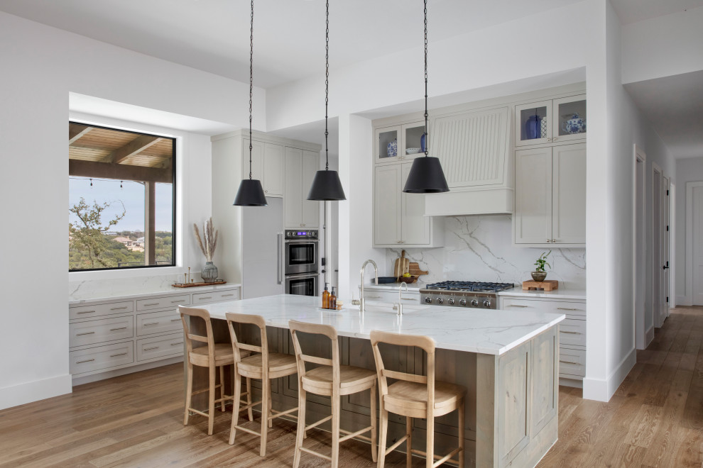 This is an example of a large country l-shaped open plan kitchen in Austin with an undermount sink, shaker cabinets, beige cabinets, quartzite benchtops, white splashback, stone slab splashback, stainless steel appliances, light hardwood floors, with island and white benchtop.