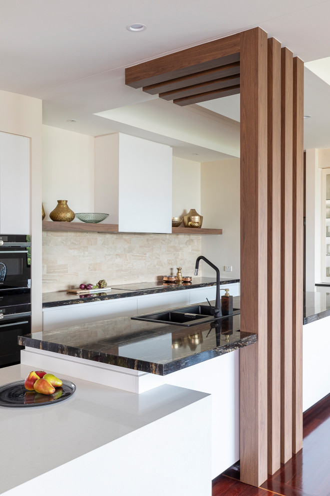 Inspiration for a large contemporary galley open plan kitchen in Brisbane with a drop-in sink, flat-panel cabinets, white cabinets, granite benchtops, beige splashback, stone tile splashback, black appliances, medium hardwood floors, brown floor, black benchtop and a peninsula.