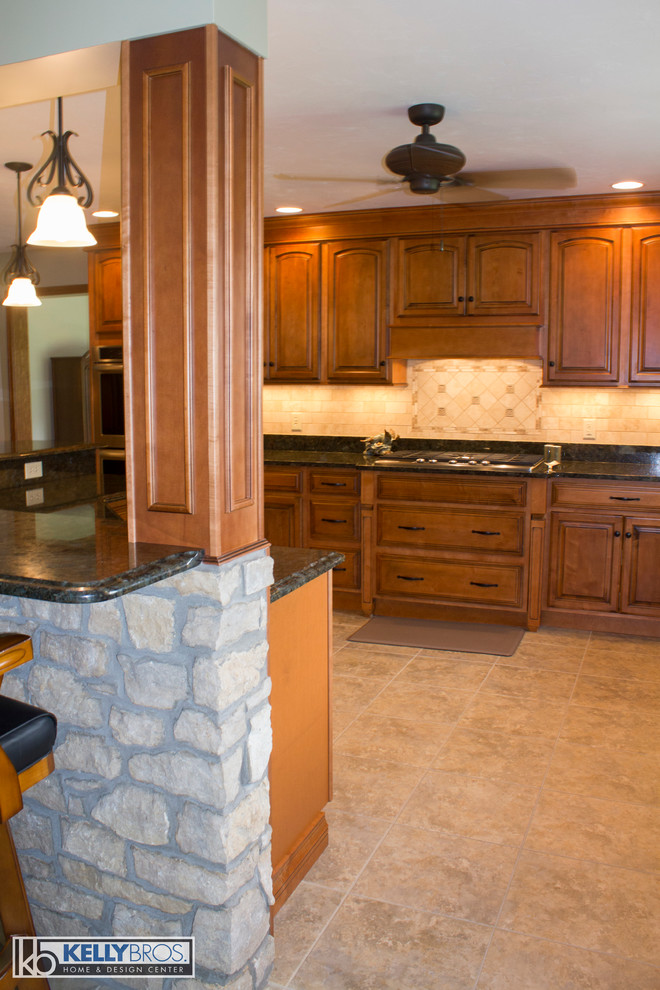 Large country l-shaped eat-in kitchen in Cincinnati with raised-panel cabinets, medium wood cabinets, granite benchtops, beige splashback, stone tile splashback, stainless steel appliances and with island.