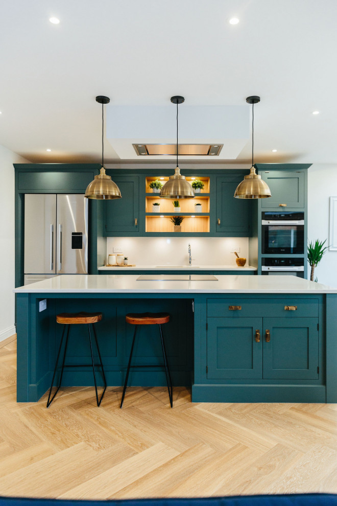 This is an example of a large contemporary l-shaped open plan kitchen in London with a built-in sink, shaker cabinets, blue cabinets, white splashback, black appliances, light hardwood flooring, an island, beige floors, white worktops and feature lighting.