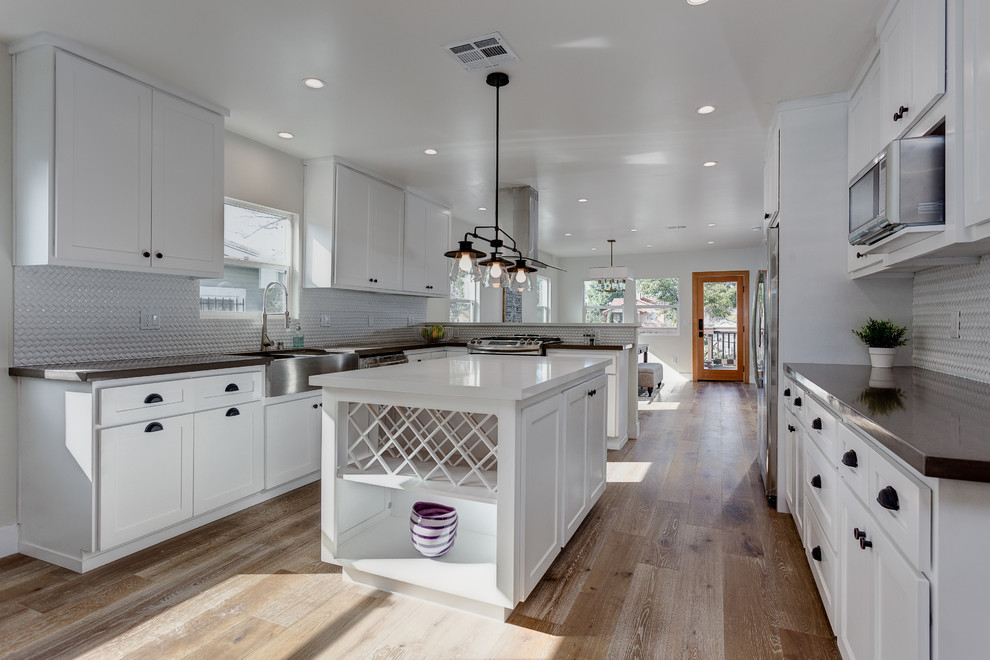 Mid-sized contemporary u-shaped open plan kitchen in Los Angeles with a double-bowl sink, recessed-panel cabinets, white cabinets, granite benchtops, white splashback, ceramic splashback, stainless steel appliances, light hardwood floors and with island.