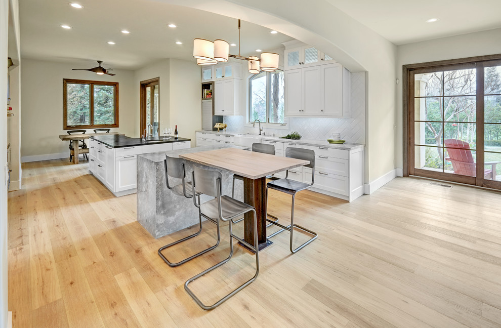 Large eclectic u-shaped open plan kitchen in Sacramento with a farmhouse sink, recessed-panel cabinets, white cabinets, marble benchtops, white splashback, porcelain splashback, stainless steel appliances, light hardwood floors and multiple islands.