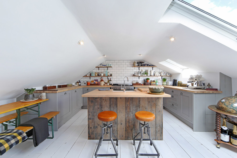 Small industrial u-shaped eat-in kitchen in London with a farmhouse sink, grey cabinets, wood benchtops, stainless steel appliances, painted wood floors, with island, raised-panel cabinets, white splashback and subway tile splashback.