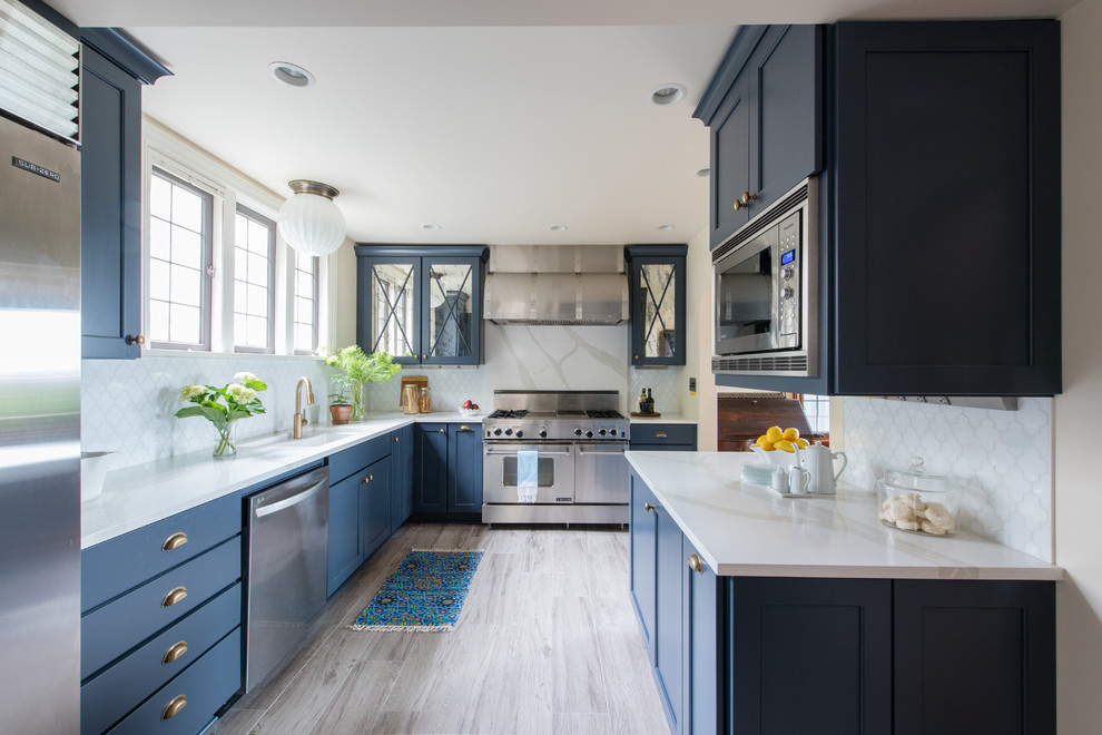 This is an example of a transitional u-shaped kitchen in Seattle with an undermount sink, shaker cabinets, blue cabinets, white splashback, mosaic tile splashback, stainless steel appliances, grey floor and white benchtop.