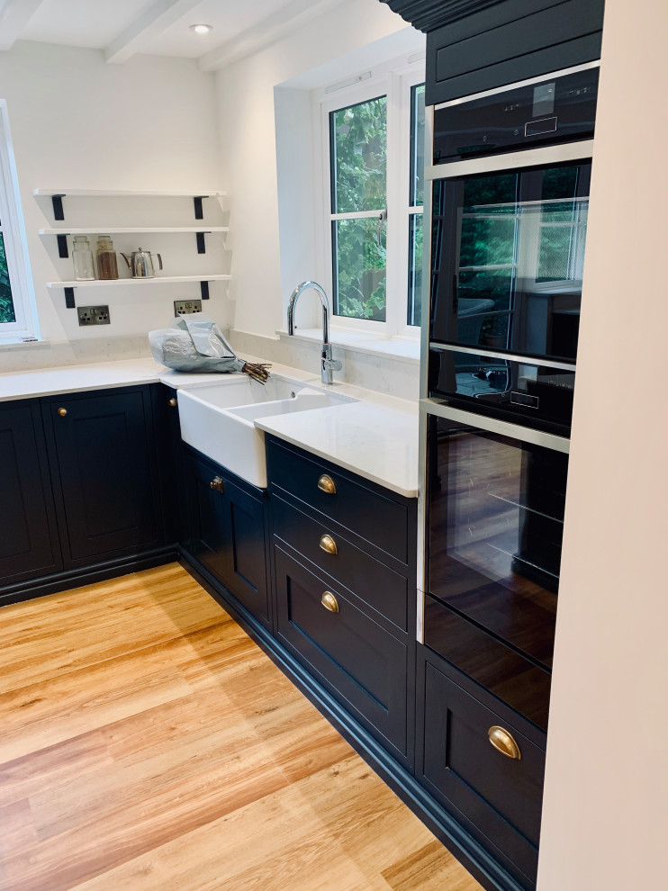 Photo of a large contemporary l-shaped eat-in kitchen in Other with a farmhouse sink, shaker cabinets, blue cabinets, quartzite benchtops, black appliances, medium hardwood floors, with island, blue floor and white benchtop.