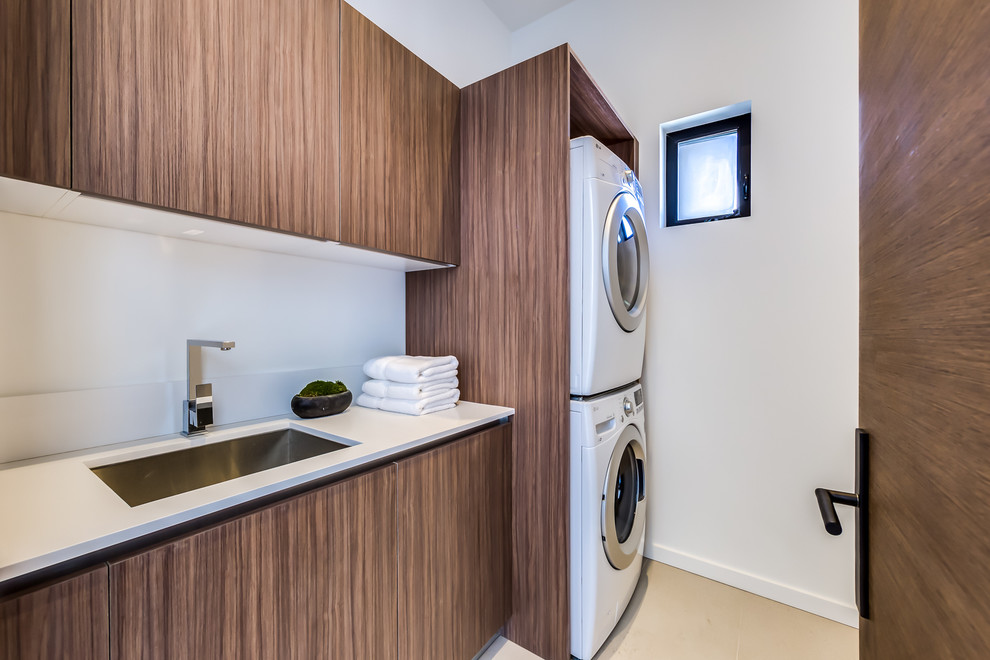 Photo of a mid-sized modern single-wall dedicated laundry room in Los Angeles with a drop-in sink, flat-panel cabinets, white walls, porcelain floors, a stacked washer and dryer and dark wood cabinets.