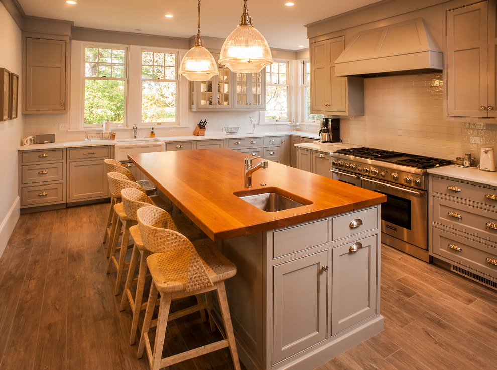 Mid-sized beach style l-shaped eat-in kitchen in Portland Maine with a farmhouse sink, shaker cabinets, grey cabinets, marble benchtops, white splashback, cement tile splashback, stainless steel appliances, ceramic floors, with island and brown floor.