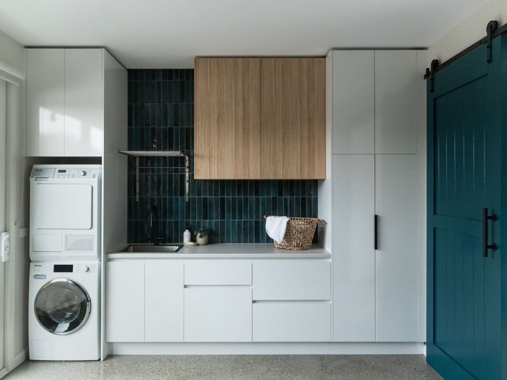 Example of a mid-sized trendy concrete floor and gray floor utility room design in Melbourne with a single-bowl sink, open cabinets, light wood cabinets, glass countertops, blue backsplash, cement tile backsplash, white walls, a stacked washer/dryer and gray countertops