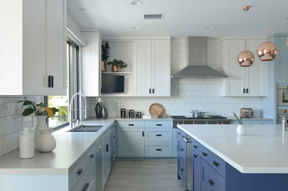 This is an example of a beach style l-shaped kitchen in Los Angeles with an undermount sink, shaker cabinets, blue cabinets, white splashback, stainless steel appliances, light hardwood floors, with island, beige floor and white benchtop.
