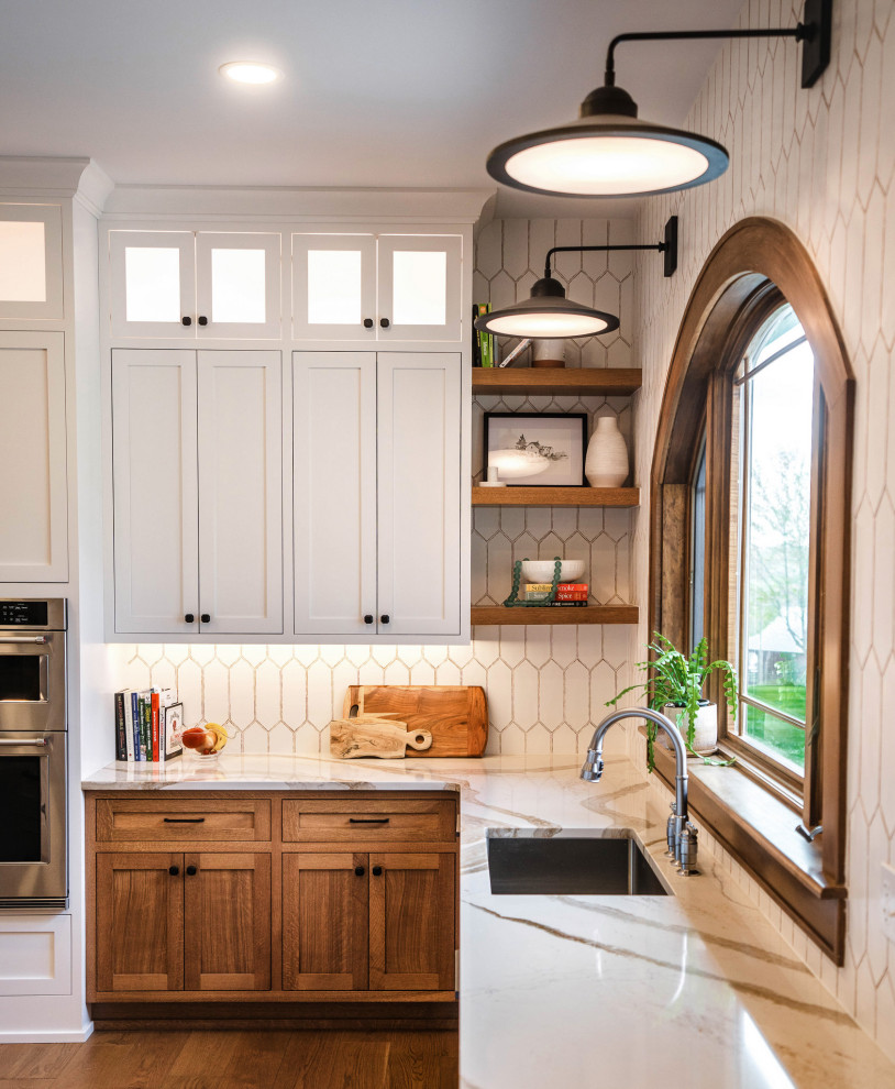 Example of a large transitional u-shaped light wood floor and brown floor eat-in kitchen design with an undermount sink, shaker cabinets, light wood cabinets, quartz countertops, white backsplash, ceramic backsplash, stainless steel appliances, an island and white countertops