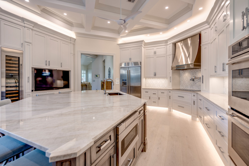 Open concept kitchen - transitional u-shaped light wood floor, beige floor and coffered ceiling open concept kitchen idea with an undermount sink, beaded inset cabinets, white cabinets, quartzite countertops, white backsplash, stone tile backsplash, stainless steel appliances, an island and gray countertops