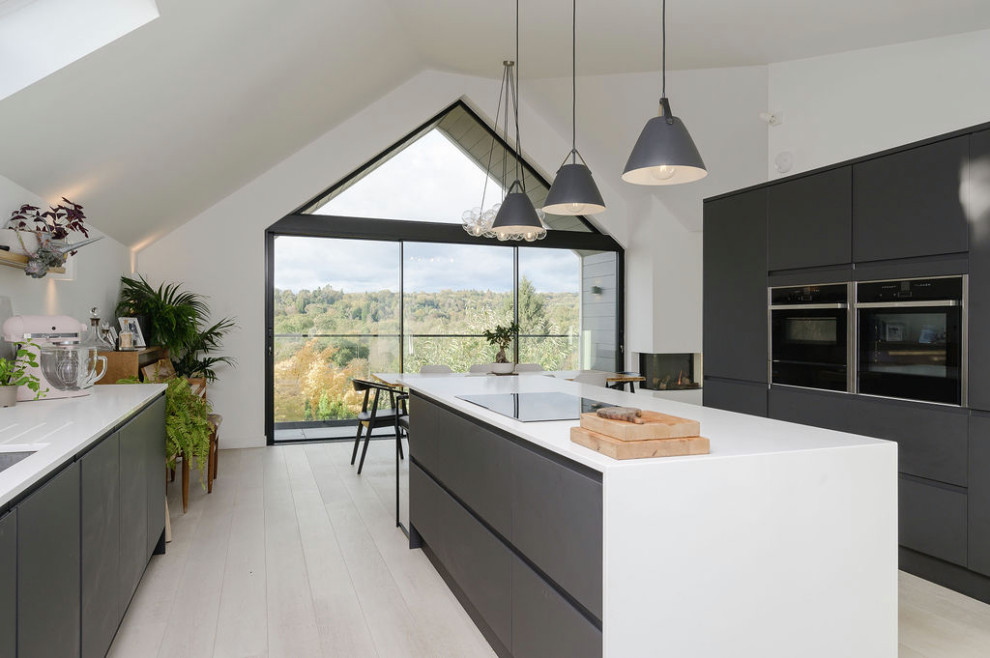 Inspiration for a large contemporary open plan kitchen with an integrated sink, flat-panel cabinets, black cabinets, solid surface benchtops, grey splashback, ceramic splashback, black appliances, light hardwood floors, with island, white benchtop and vaulted.