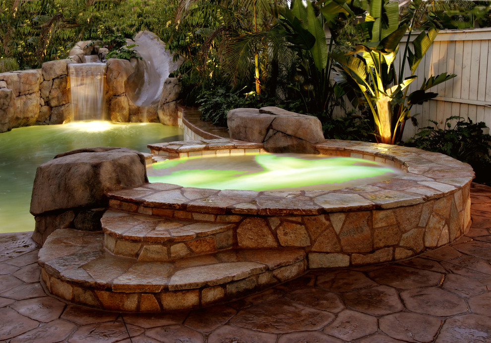 Design ideas for a mid-sized tropical backyard custom-shaped pool in Orange County with natural stone pavers and a hot tub.
