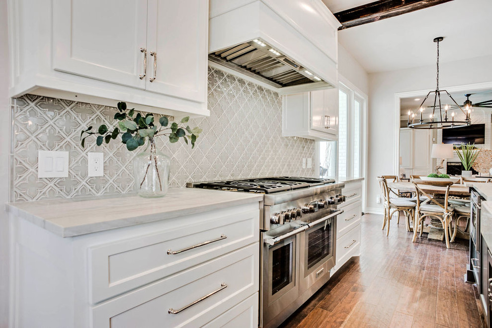 Eat-in kitchen - mid-sized traditional u-shaped medium tone wood floor and brown floor eat-in kitchen idea in Dallas with a farmhouse sink, recessed-panel cabinets, white cabinets, quartzite countertops, gray backsplash, ceramic backsplash, paneled appliances, an island and white countertops