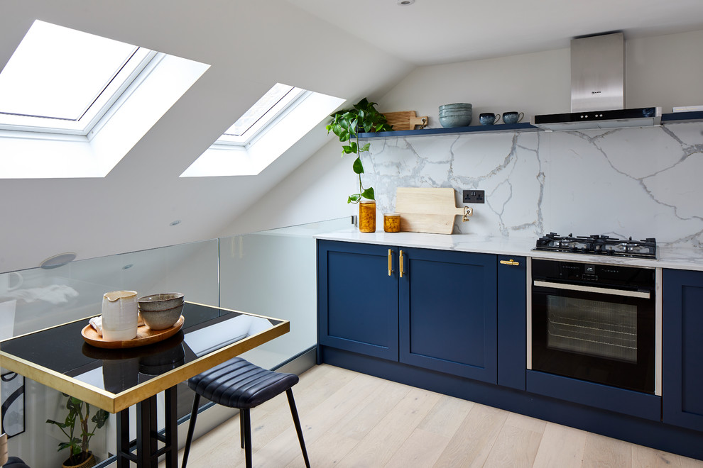 This is an example of a contemporary kitchen in London with shaker cabinets, blue cabinets, white splashback, stone slab splashback, black appliances, light hardwood floors, beige floor and white benchtop.