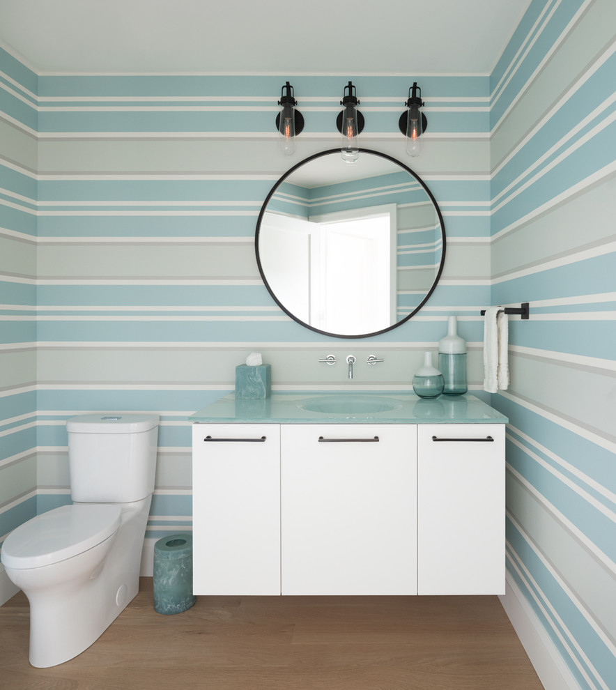 Beach style powder room in Minneapolis with flat-panel cabinets, white cabinets, a two-piece toilet, multi-coloured walls, light hardwood floors, an integrated sink, glass benchtops and blue benchtops.