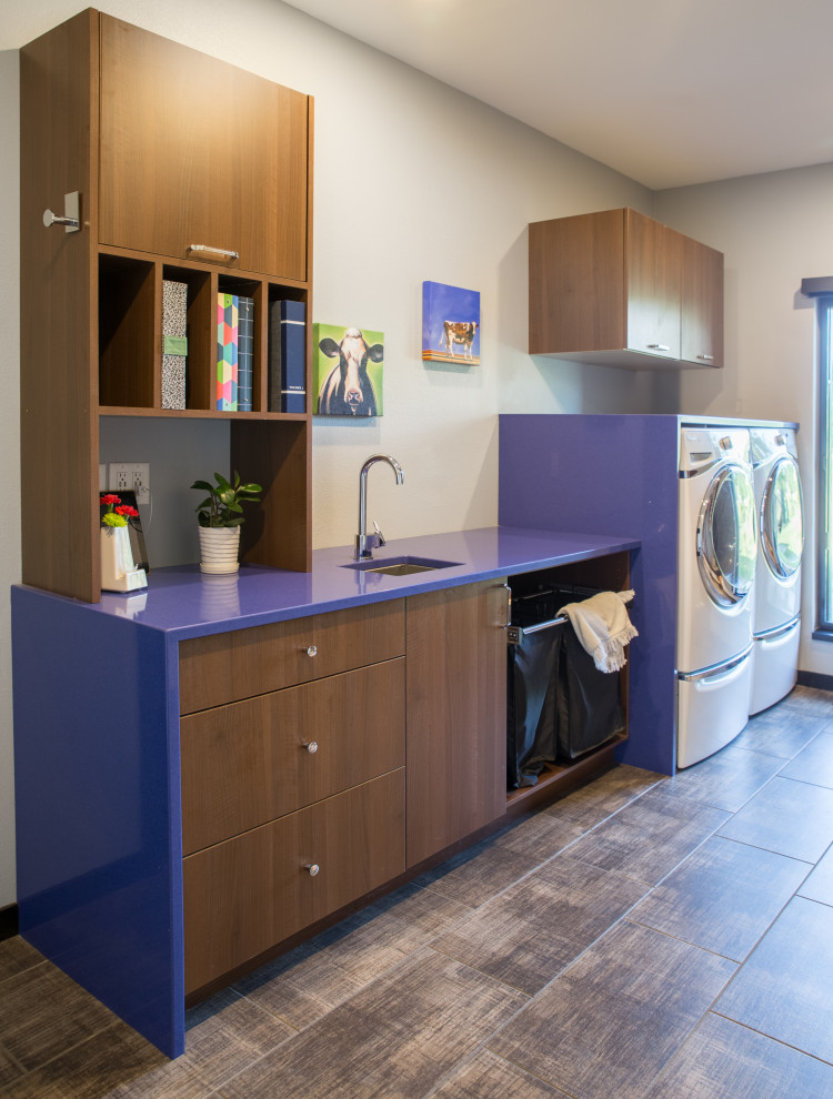 Idéer för att renovera en mellanstor funkis blå linjär blått tvättstuga enbart för tvätt, med en undermonterad diskho, släta luckor, skåp i mellenmörkt trä, bänkskiva i kvarts, vita väggar, en tvättmaskin och torktumlare bredvid varandra, skiffergolv och brunt golv