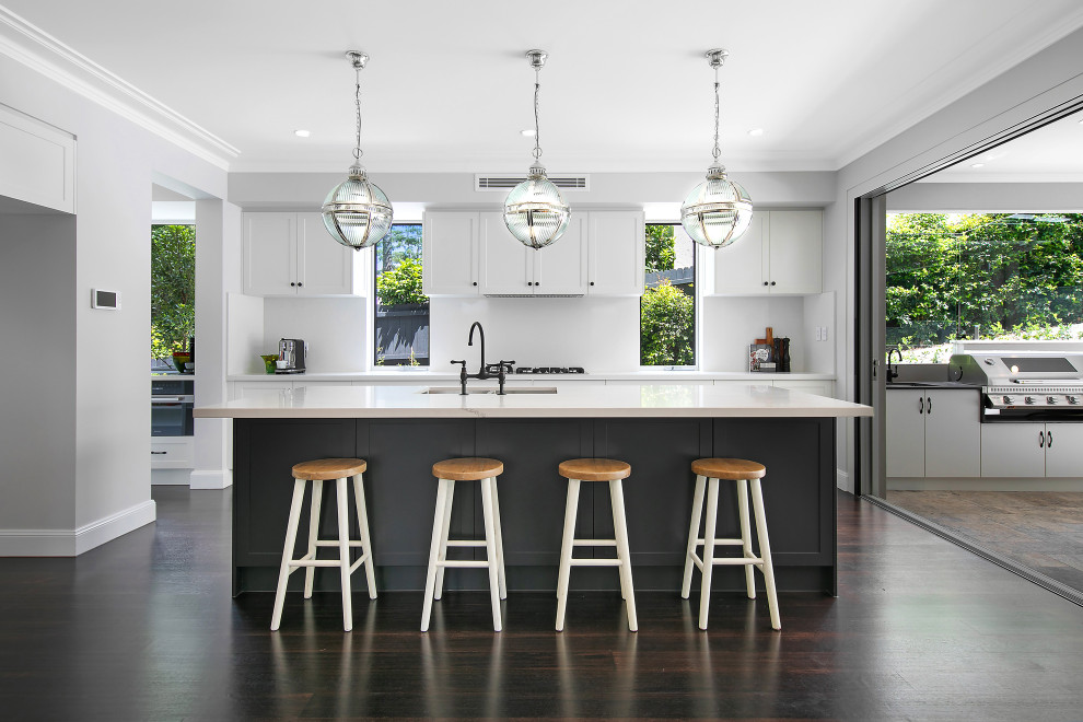 Photo of a large beach style galley eat-in kitchen in Sydney with a double-bowl sink, shaker cabinets, white cabinets, white splashback, glass sheet splashback, black appliances, dark hardwood floors, multiple islands, black floor and white benchtop.