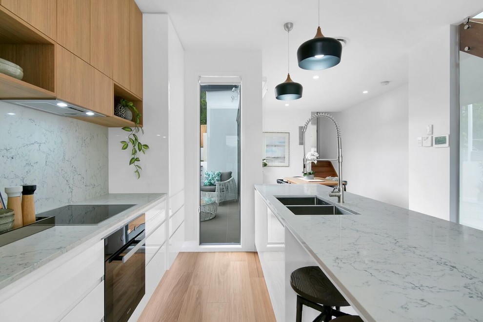 Photo of a contemporary galley separate kitchen in Brisbane with an undermount sink, flat-panel cabinets, medium wood cabinets, grey splashback, black appliances, light hardwood floors, with island, beige floor and grey benchtop.
