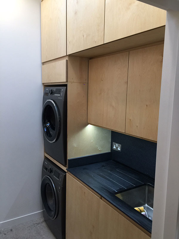 Inspiration for a mid-sized eclectic l-shaped laundry room in Dublin with an integrated sink, flat-panel cabinets, light wood cabinets, quartzite benchtops, black splashback, slate splashback, concrete floors, grey floor and black benchtop.